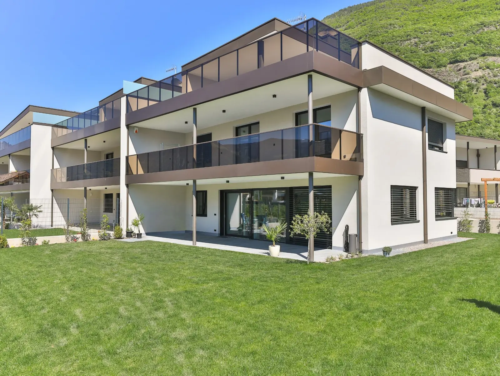 Residence Floris Gargazon — Gartenansicht mit umlaufenden Balkonen und Dachterrasse vor bewaldetem Hang