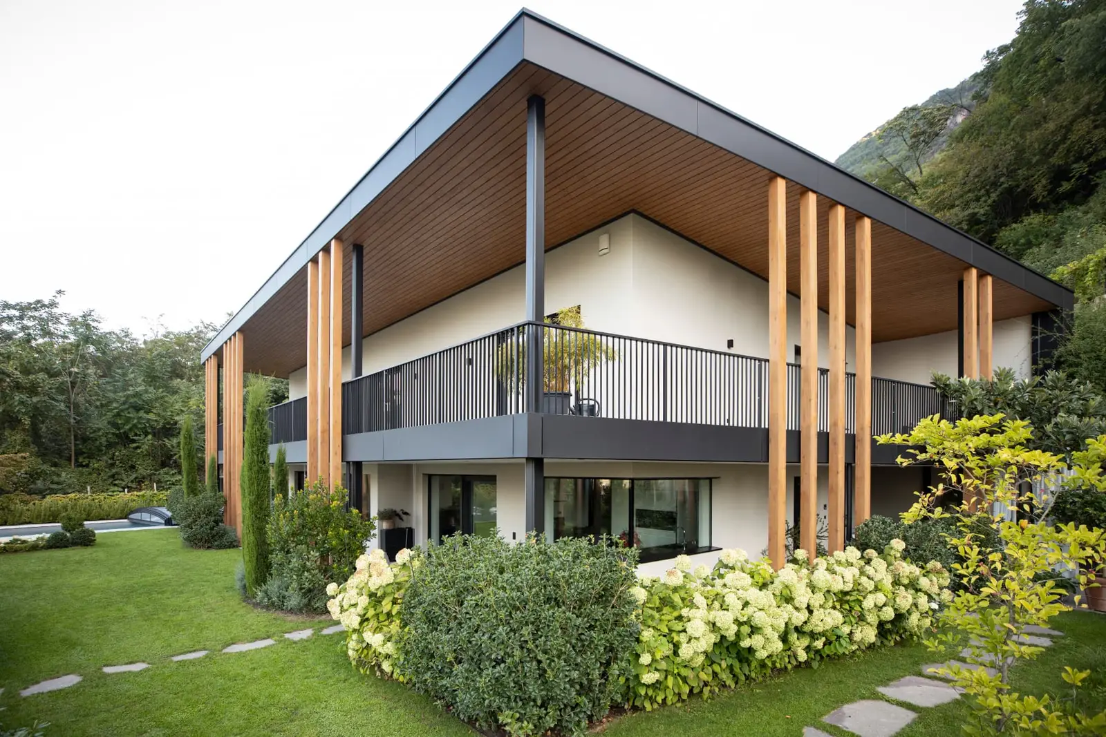 Villa F Burgstall — Gebäudeansicht mit Holzstützen, umlaufendem Balkon und Gartenanlage
